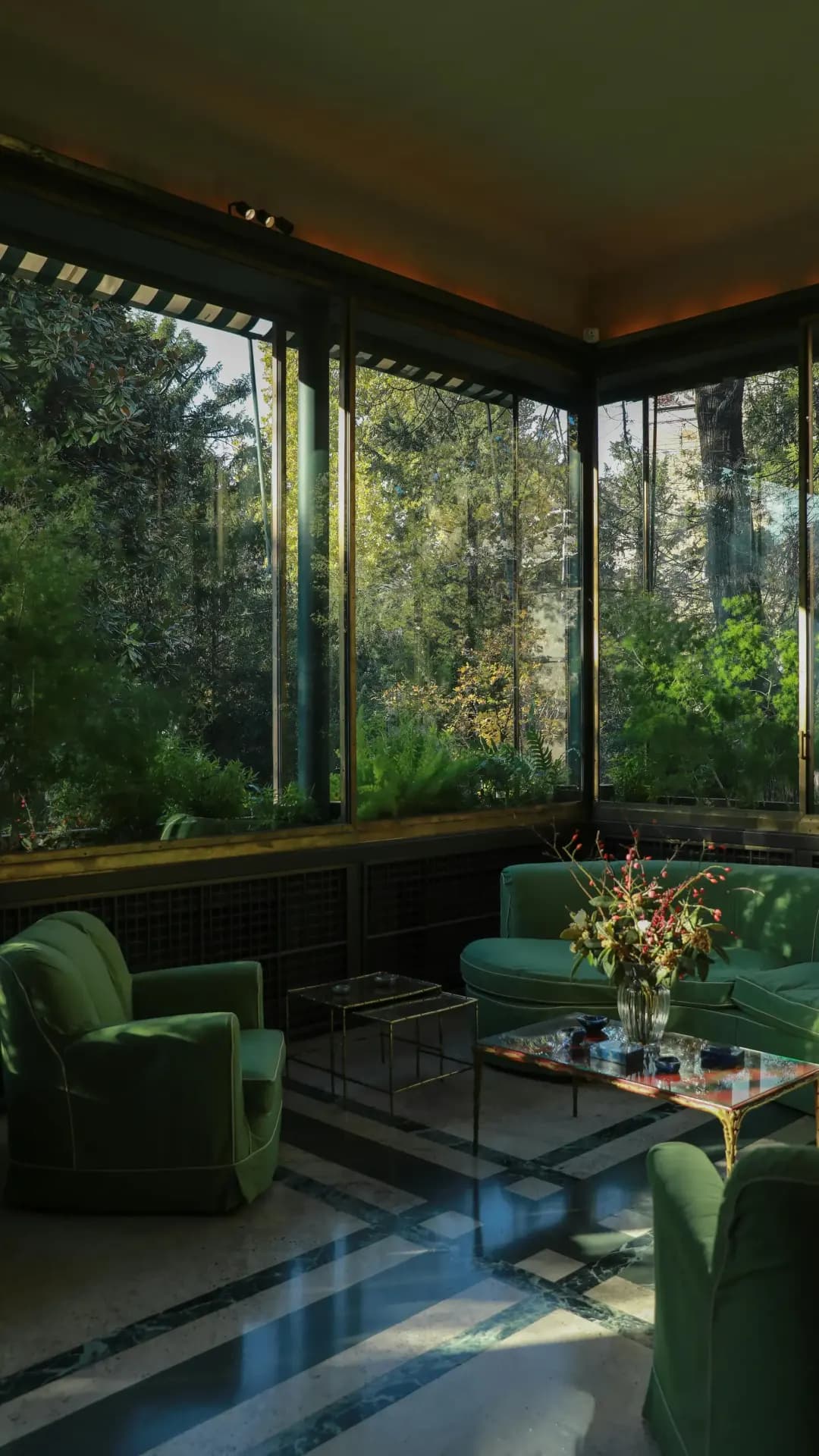 Glass-enclosed sunroom with green velvet seating and lush garden views