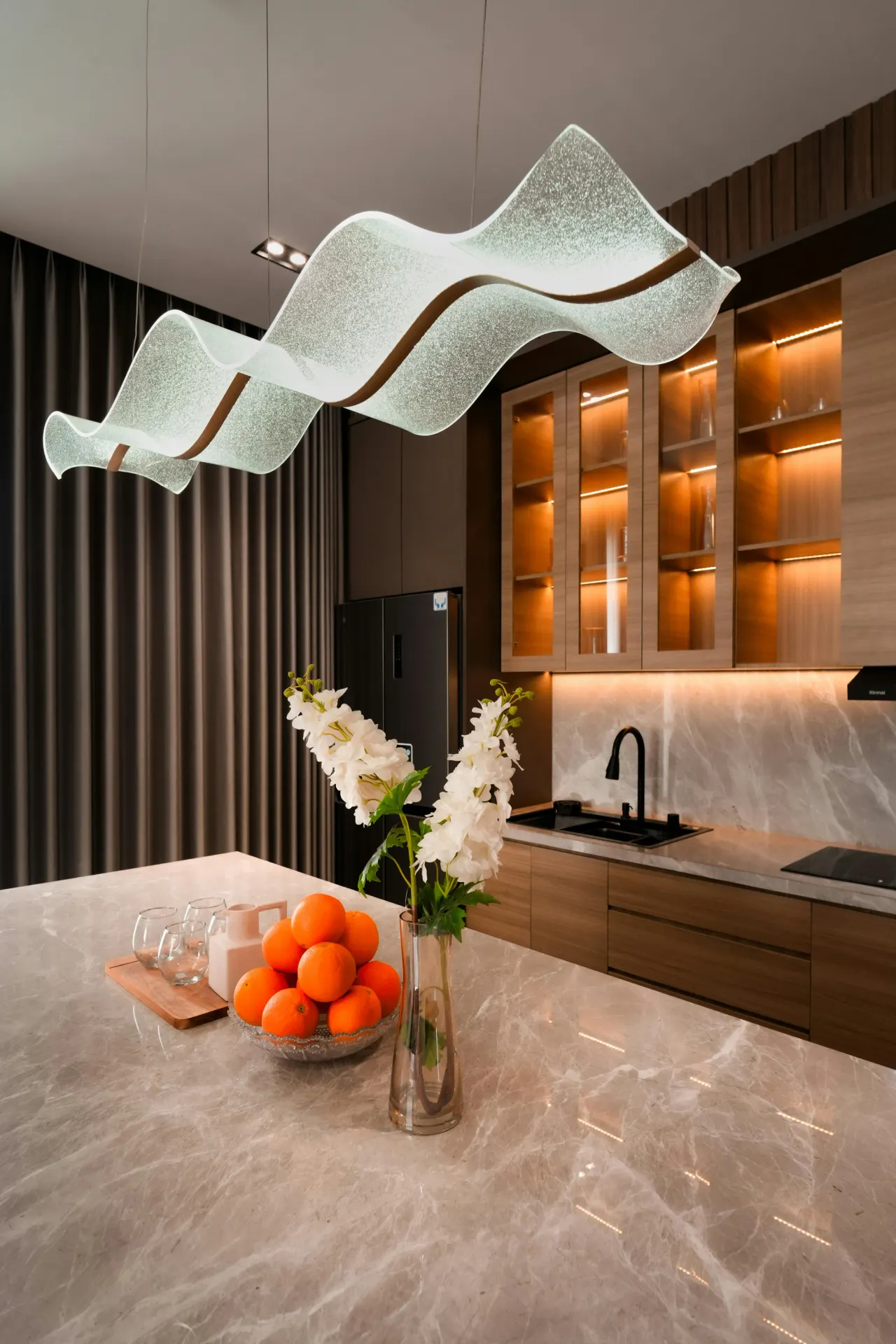 Modern luxury kitchen featuring dramatic sculptural wave pendant light over marble island with warm wood cabinetry
